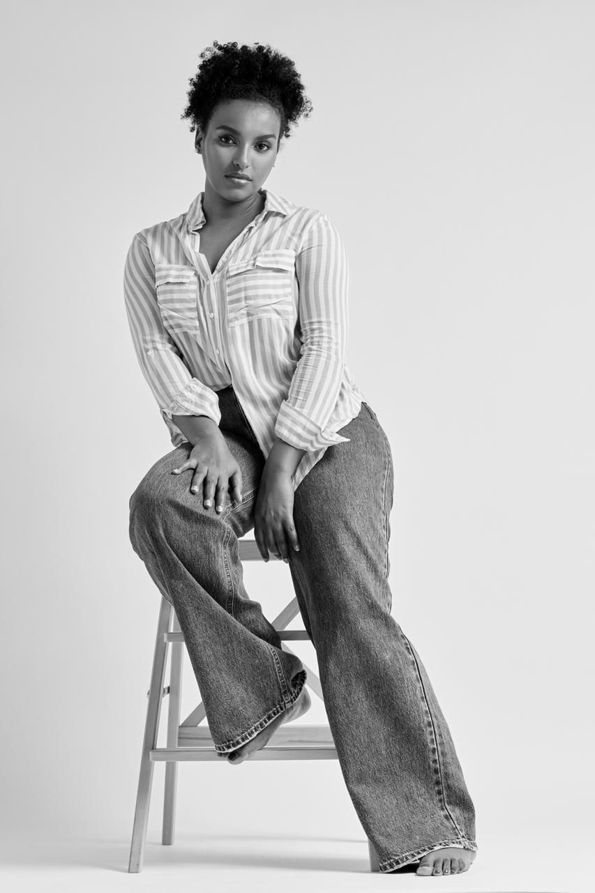 Person posing on a high stool, wearing a striped shirt and wide-leg jeans.